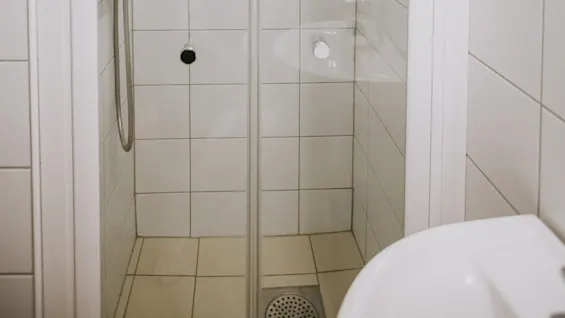 En brusekabine med glasdøre og en bruserhoved er omgivet af et flisebelagt, minimalistisk badeværelsesmiljø.