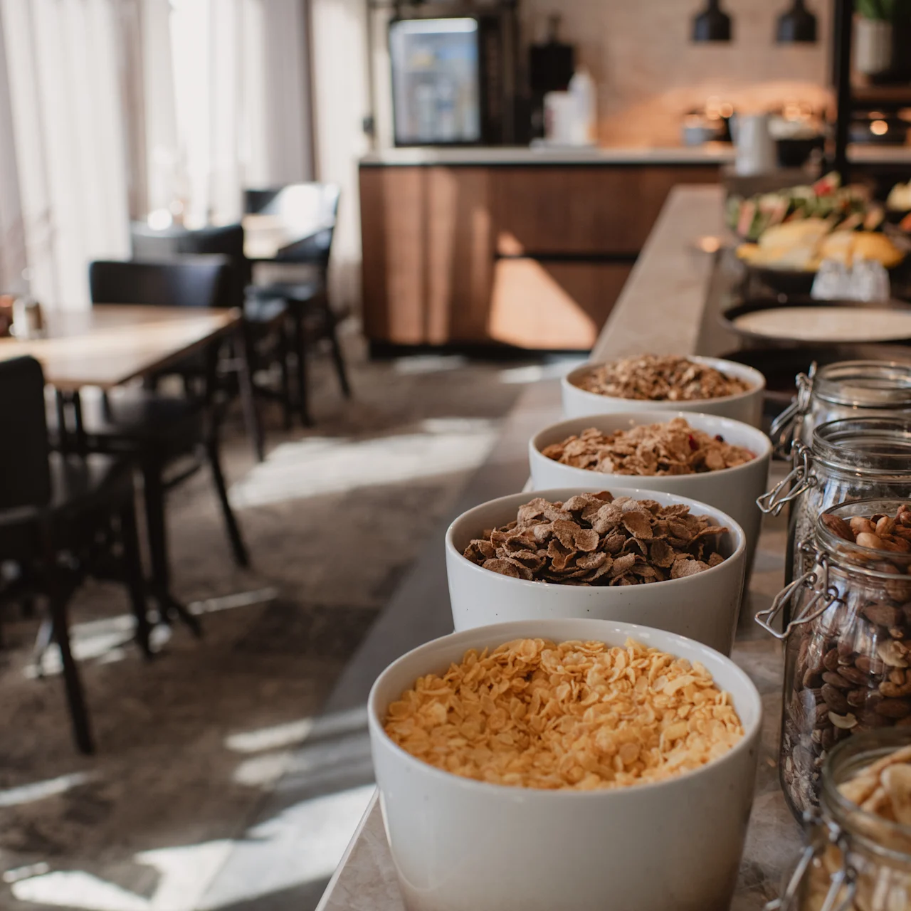 Bowls and jars of assorted cereals and nuts are displayed on a countertop, set in a cozy dining area with tables and soft lighting, creating a warm, inviting breakfast atmosphere.