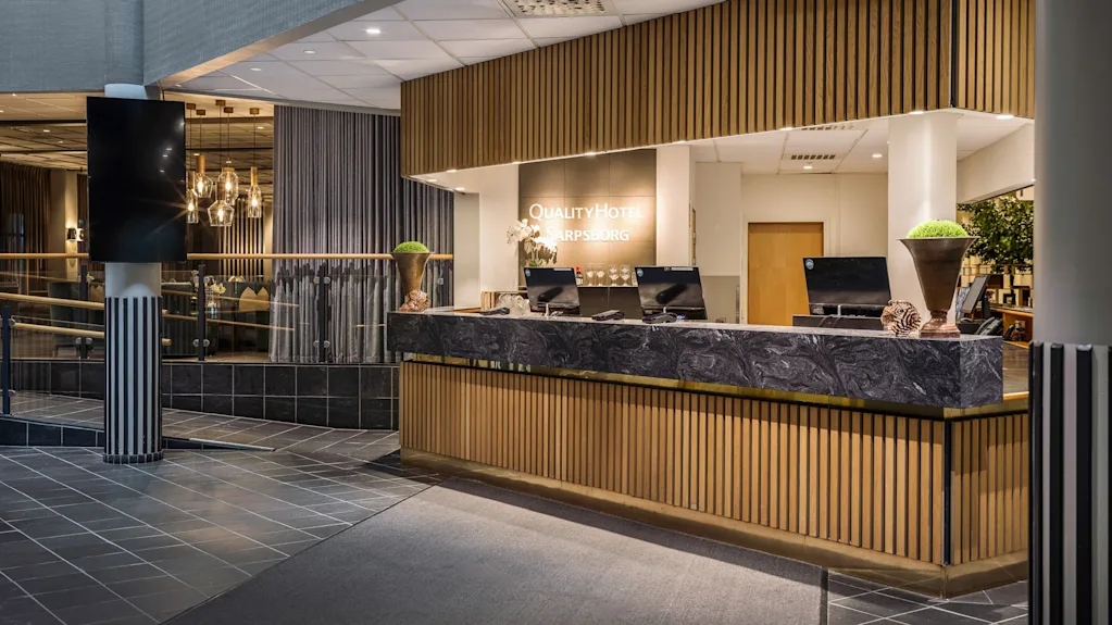 Reception med datorer och dekorationer står i en modern hotellobby. Bakom skrivbordet står en skylt med texten "QUALITY HOTEL SARPSBORG" med stilfull belysning och kaklat golv.