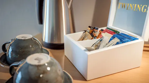A white box filled with various tea packets labeled "TWININGS" is open on a wooden table next to two inverted ceramic cups and a stainless steel electric kettle.