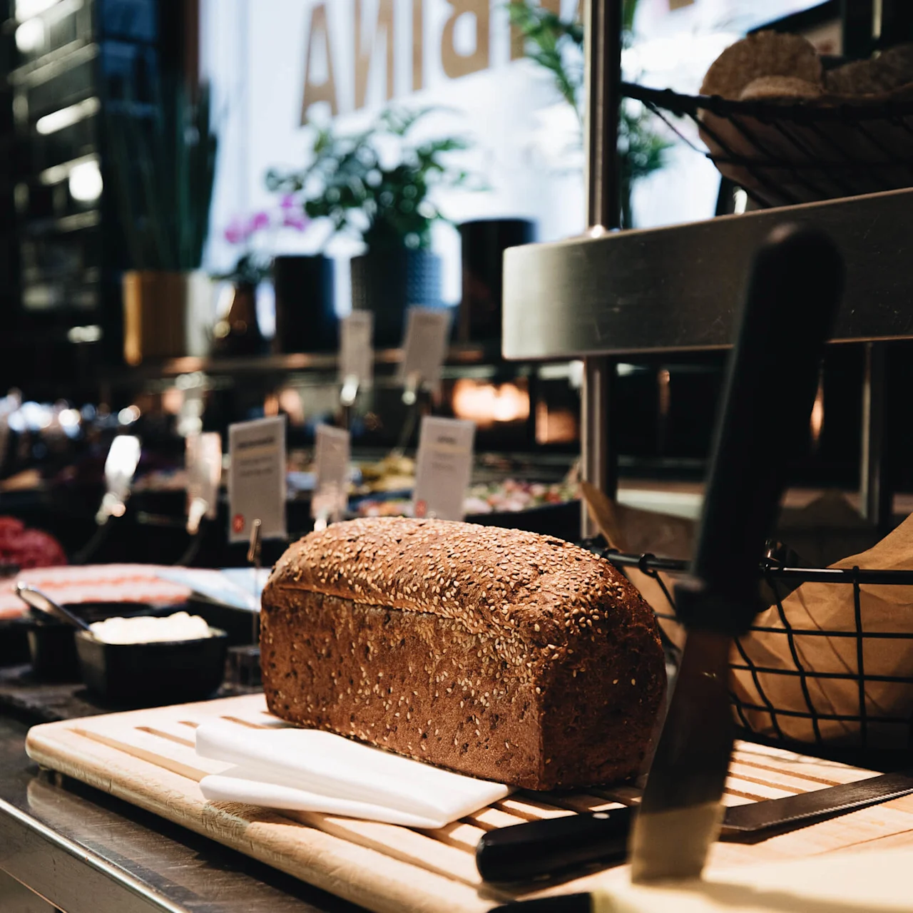 A seeded loaf rests on a wooden cutting board beside a butter slab and knife in a buffet setting with plants and labeled dishes in the background. Text: "ARIA."