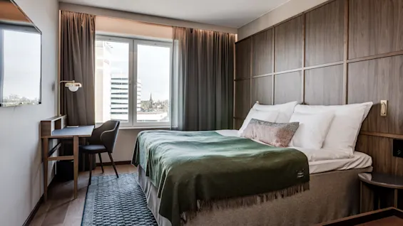 En seng med grønt tæppe og hvide puder er pænt arrangeret i et hotelværelse med trævægge. I nærheden står et skrivebord med en stol ved et stort vindue.