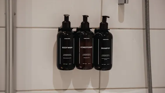 Kolme tummaa pulloa, joissa on merkinnät Body Wash, Conditioner ja Shampoo, on kiinnitetty valkoiseen kaakeloituun suihkuseinään.