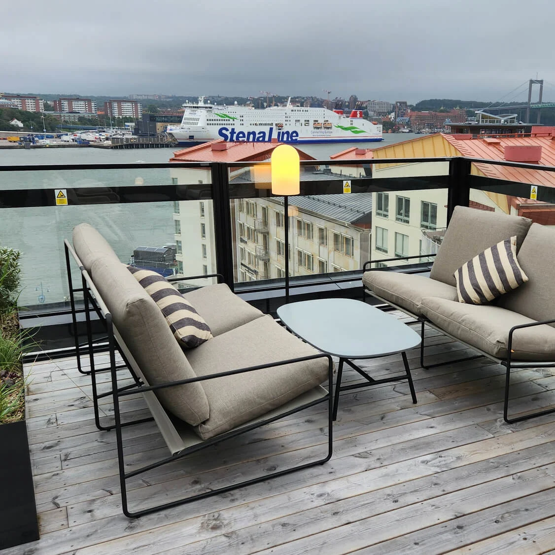 Outdoor seating area on a wooden deck features two cushioned chairs with striped pillows and a small table. The background displays a harbor with a ship labeled "Stena Line" and an industrial crane labeled "ERIKSBERG."
