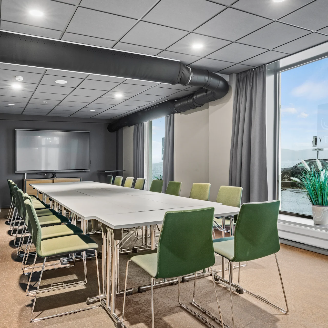 Konferencelokale med rektangulært bord omgivet af grønne stole, med flipover og whiteboard mod en grå væg. Store vinduer viser en udendørsscene, med en plante i nærheden.