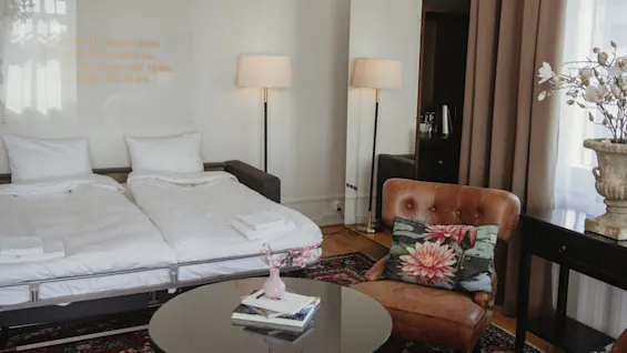 En pænt redt seng med hvidt sengetøj i et hotelværelse, omgivet af en brun læderstol, et rundt sofabord med bøger, gulvlamper og dekorative blomster.