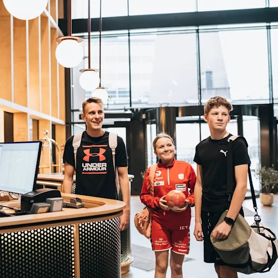 Personer smiler i en moderne hotellobby, én ved receptionen og tre andre, der træder ind, iført sportstøj.