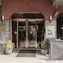 Ingången av ett hotell med glasdörrar och krukväxter, omgiven av en röd och grå fasad. Text på dörrarna: "PLAZA HOME HOTEL".
