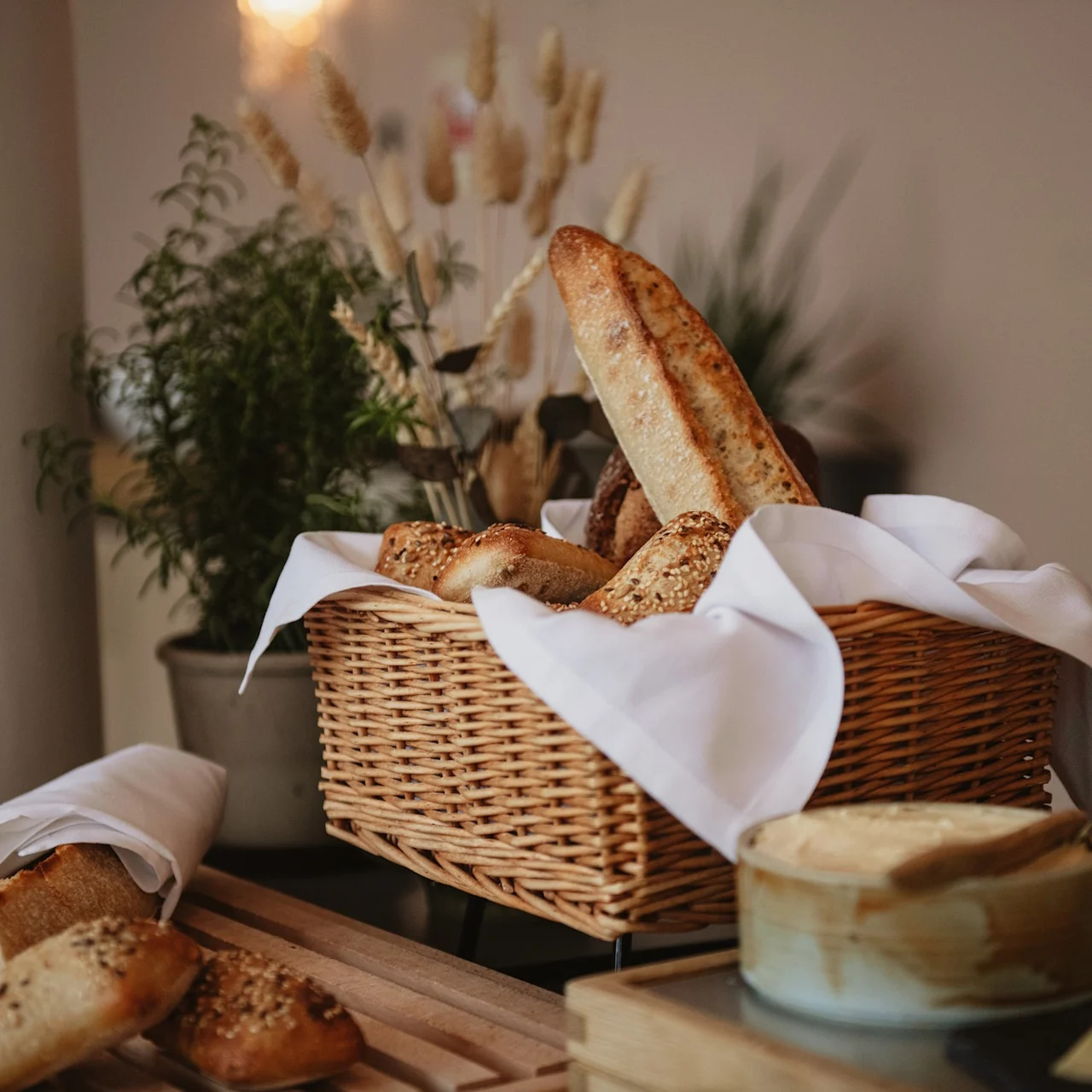 A wicker basket holds various loaves of bread, covered with a white cloth, on a wooden table surrounded by potted plants in a softly lit room.