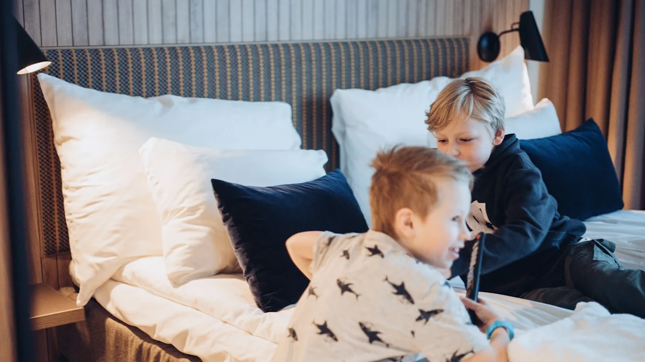 Two children sit playfully on a hotel bed, surrounded by white pillows and dark blue cushions, with warm lighting in a cozy room.