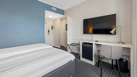 A tidy hotel room with a made bed, a mounted TV, desk, chair, and a suitcase stand. No visible text.
