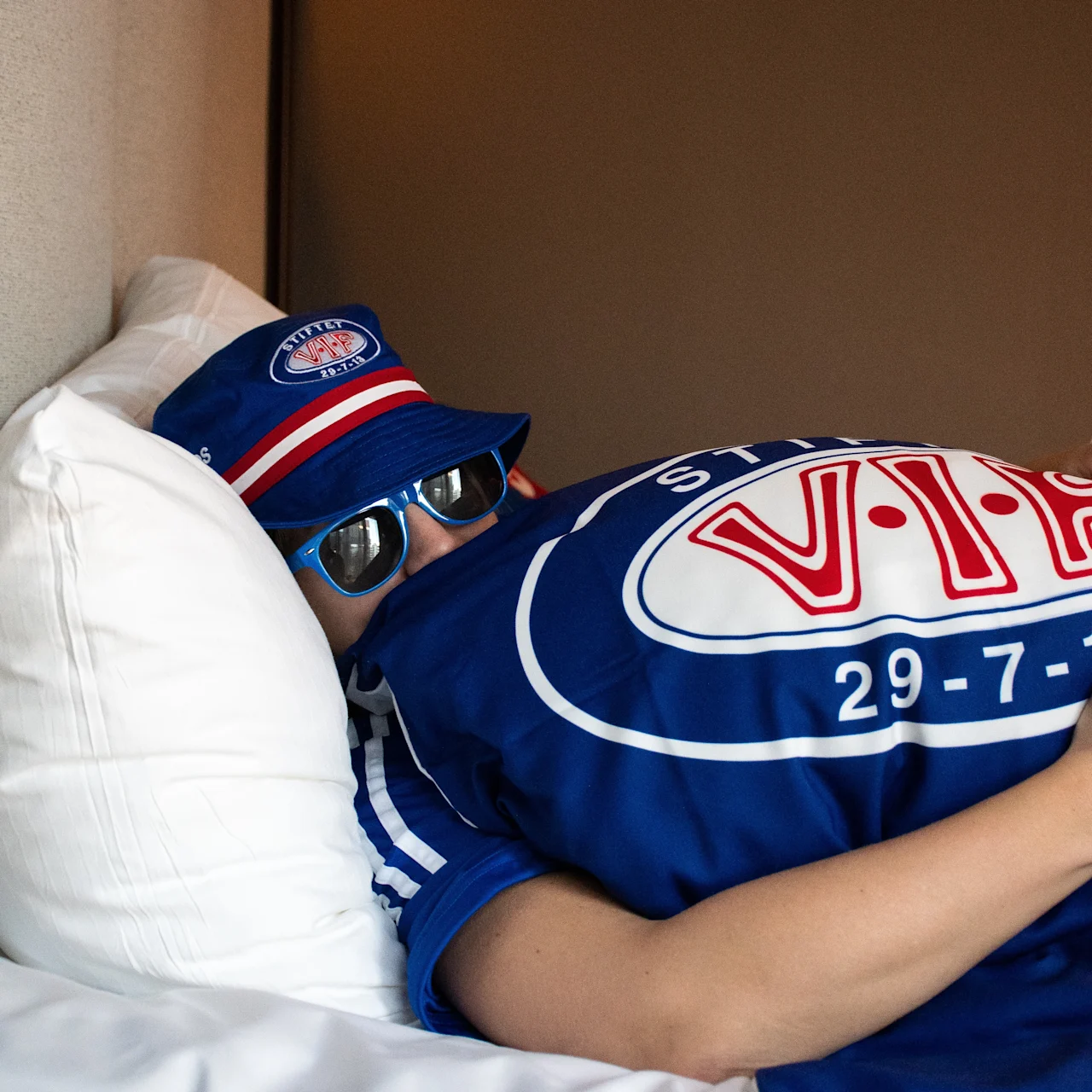 En person i blå hatt og solbriller slapper av i sengen, iført en blå skjorte. Begge plaggene har teksten VIF 29 7 13.