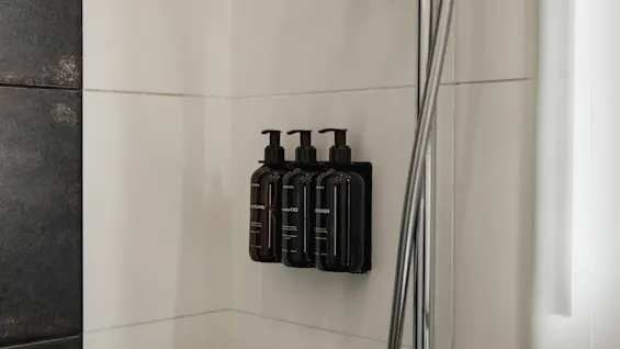 Tre pumpeflasker mærket balsam, shampoo og kropsvask monteret på en flisebelagt brusevæg.