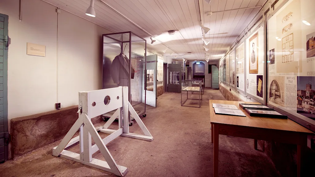 A historical museum exhibit in a long hallway with a wooden stockade, display cases, and framed documents on the wall. Visible text: BRITTISK STRAFF.