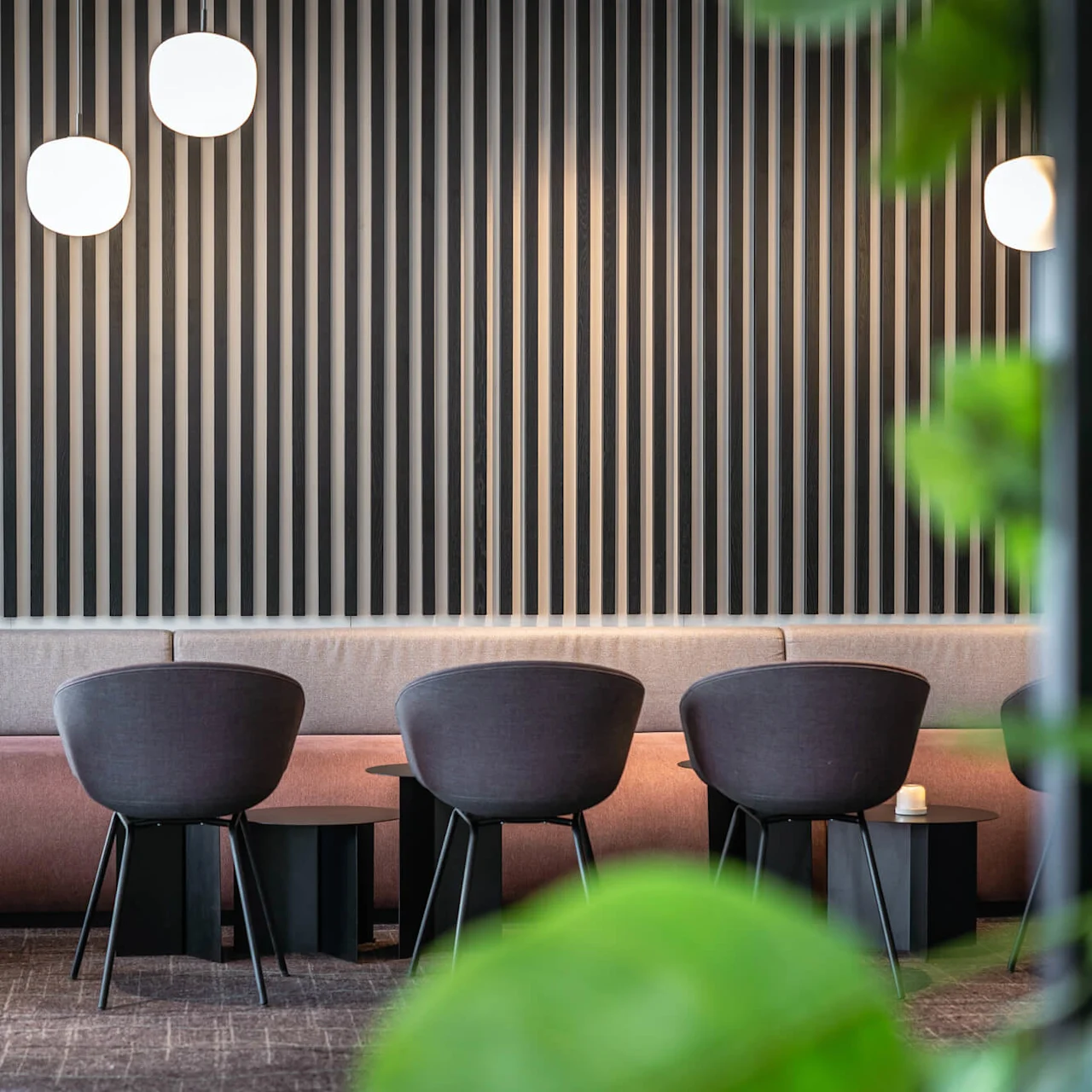 Stole er på række i et moderne loungeområde, vendt mod en stribet accentvæg. Runde lamper hænger over, med grønne planter delvist synlige i forgrunden.