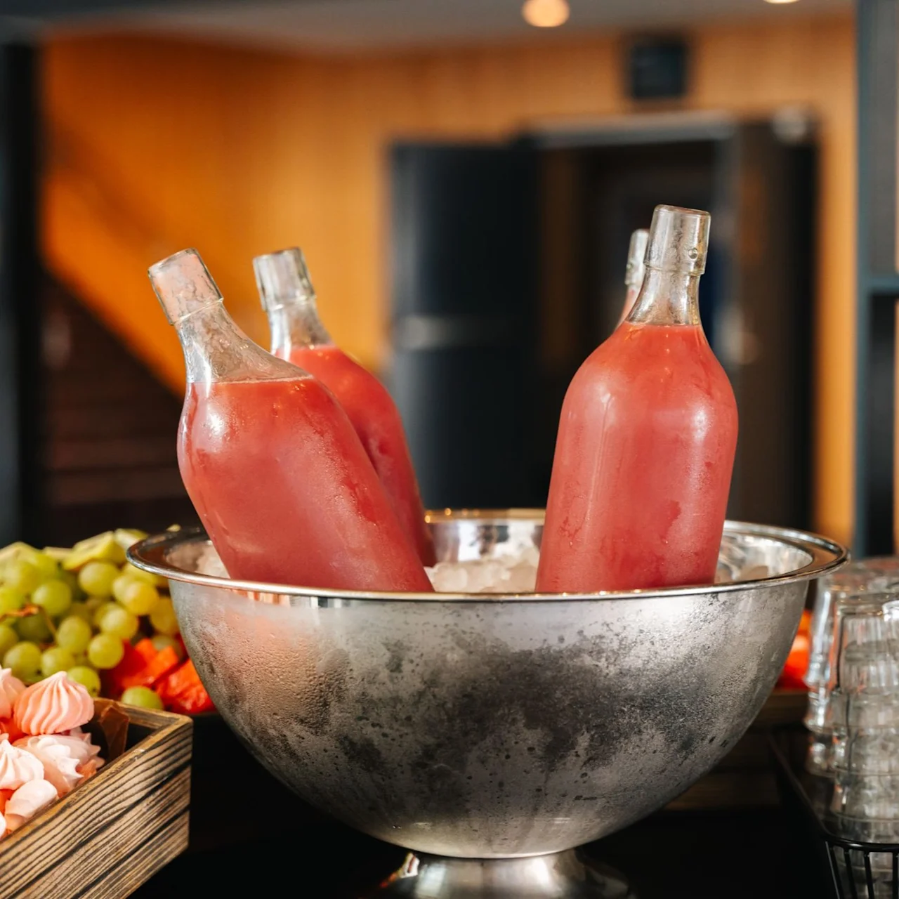 Three glass bottles filled with pink liquid stand submerged in an ice-filled metal bowl. Next to them, a wooden tray holds assorted grapes and pink meringues, with stacked glasses nearby.
