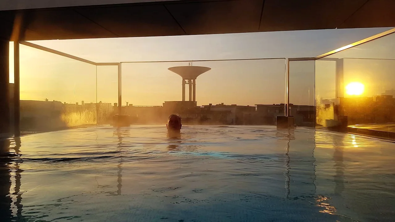 Quality Hotel™ Öresund – Rooftop Spa. En person slapper av i et dampende takbasseng med en gyllen solnedgang over byen.