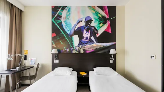 Två enkelsängar i ett hotellrum, toppade av en väggmålning av en DJ som uppträder, med ett skrivbord och ett fönster prytt med bruna gardiner.