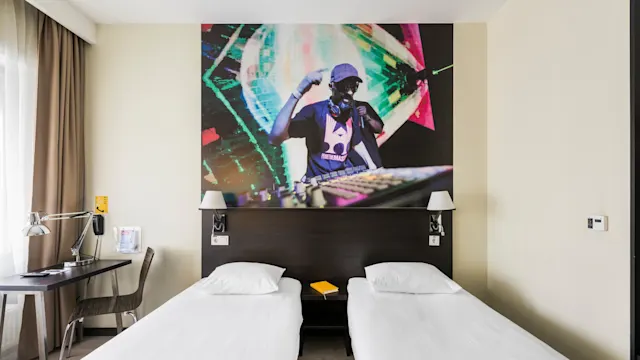 Two single beds in a hotel room, topped by a mural of a DJ performing, with a desk and a window adorned with brown curtains.