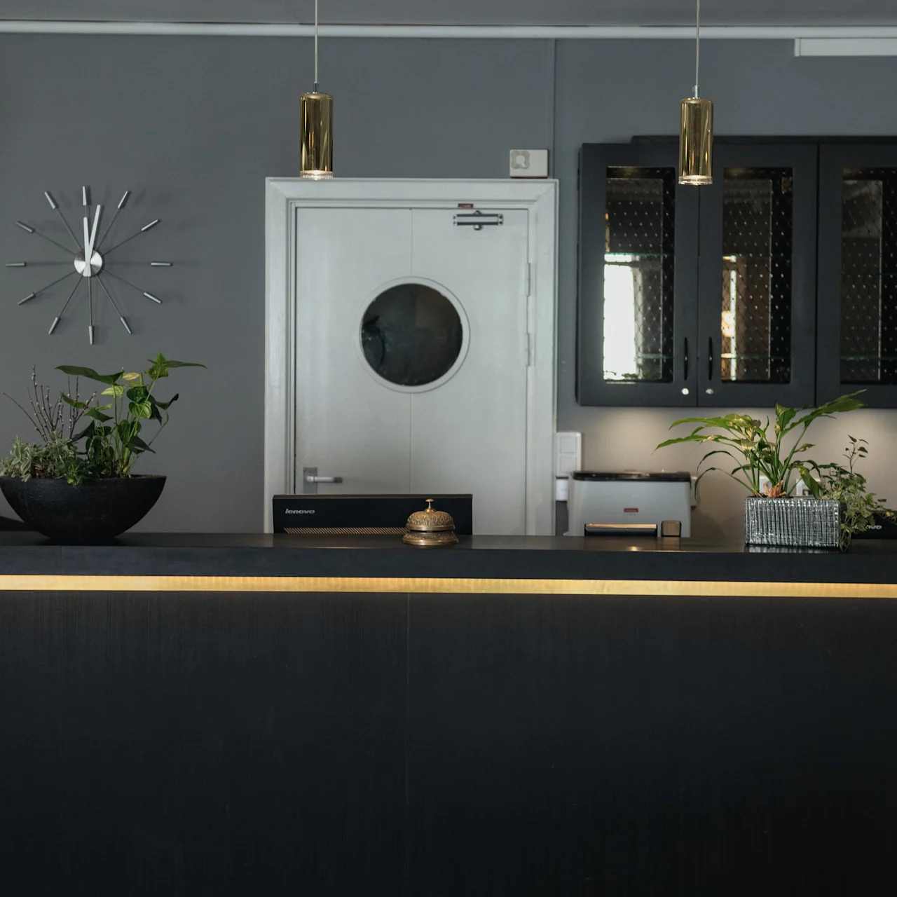 A reception desk features a small bell and plant, beneath pendant lights. Behind it, a white door with a round window and a wall clock enhance the modern, minimalist design.