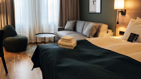 Bed with folded towels on top, set in a cozy hotel room featuring a sofa, armchair, round table, lamp, framed art, and large curtained windows.