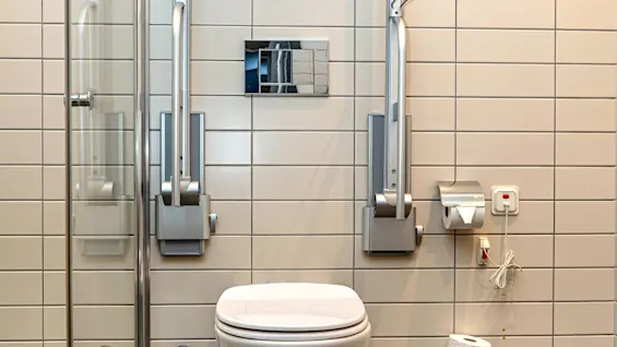 A clean, accessible bathroom with a toilet, supportive grab bars, a glass shower, and an emergency call system.