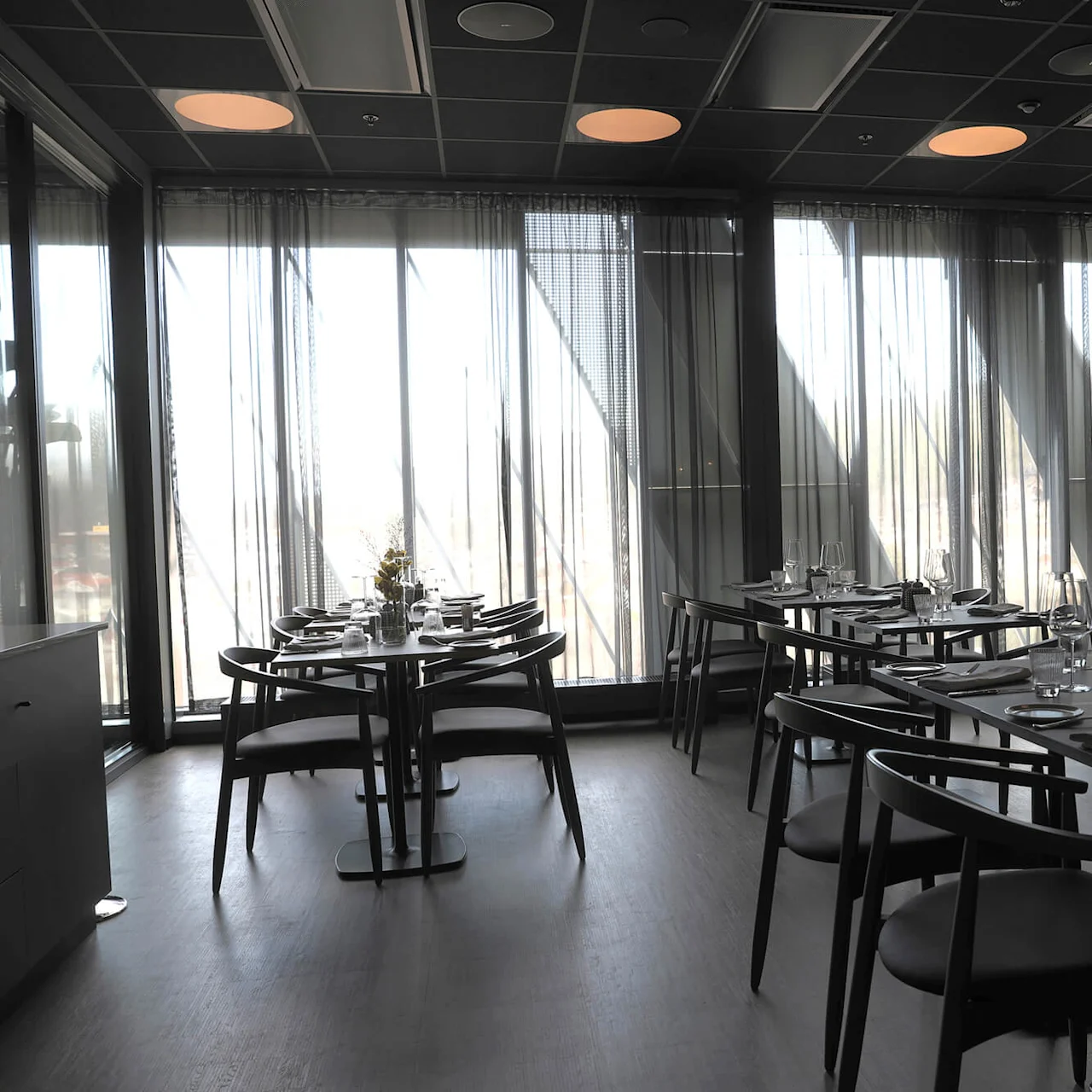 An empty modern restaurant with set tables, floor-to-ceiling windows, and stylish interior under soft lighting.
