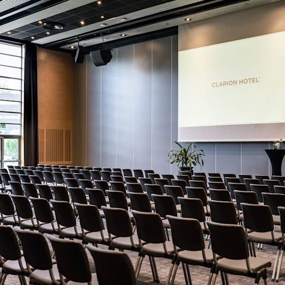 A spacious conference room with rows of chairs facing a large screen displaying CLARION HOTEL. Natural light fills the room.