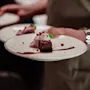 A server carries two white plates, each holding a chocolate dessert topped with berry sauce and garnished with cream and mint, in a dimly lit dining setting.