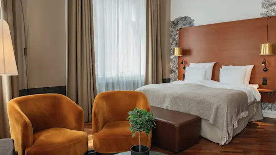Et indbydende hotelværelse med en komfortabel seng, et par plysj lænestole og blødt naturligt lys fra et vindue.