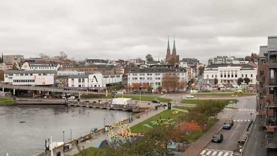 Et overskyet bylandskap med en elv, bygninger ved vannet, en park med fargerike trær, veier og en kirke med spirer.