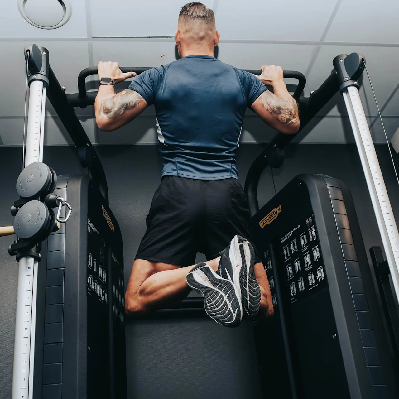 A person performs a pull-up on gym equipment, wearing athletic clothing in a fitness facility. The equipment displays "TECHNOGYM" and exercise instructions.