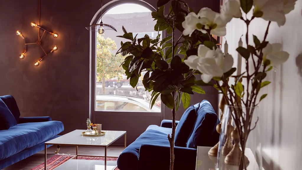 Mysig hotellounge med blå sammetssoffor, varm belysning, gröna växter och ett stort bågformat fönster.