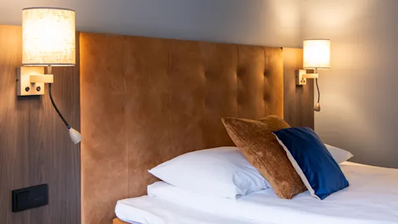 A bed with a tufted brown headboard holds white pillows and a blue cushion; wall-mounted lamps provide warm light in a modern bedroom setting.