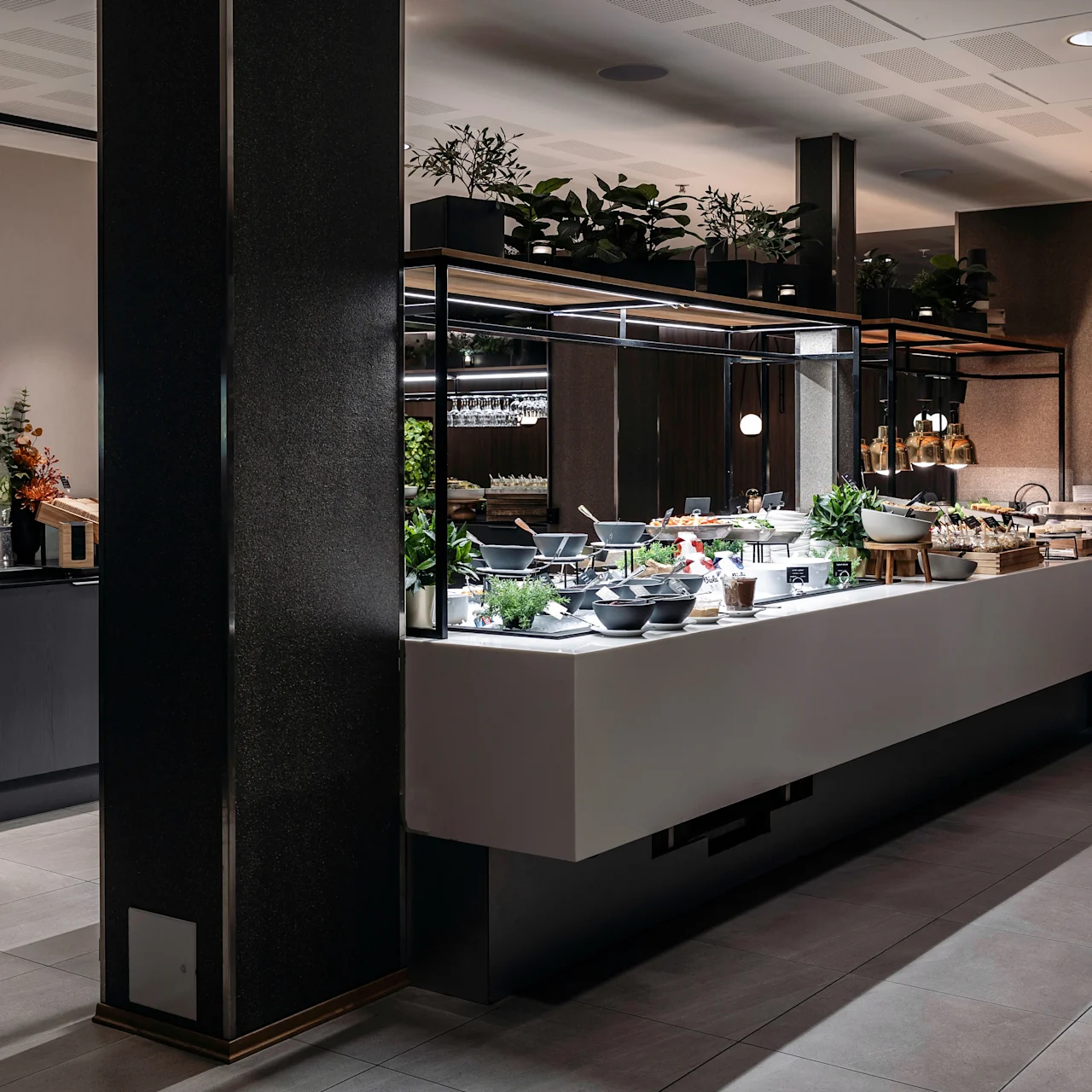 Buffet table displays assorted food and bowls under hanging lights in a modern dining room with plants and seating in the background. Various condiments and beverages are arranged on a countertop.