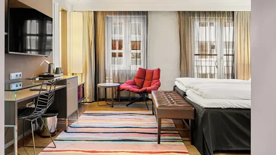 Et moderne hotelværelse med en dobbeltseng, rød lænestol, stribet tæppe og vægmonteret tv i et lyst rum med store vinduer. Tekst: Ingen.
