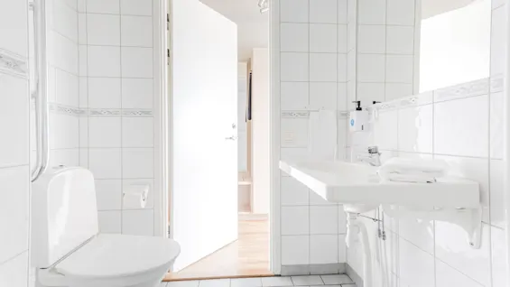 A bright, clean bathroom with white tiles, a toilet, and a sink with a mirror. An open door leads to another room.
