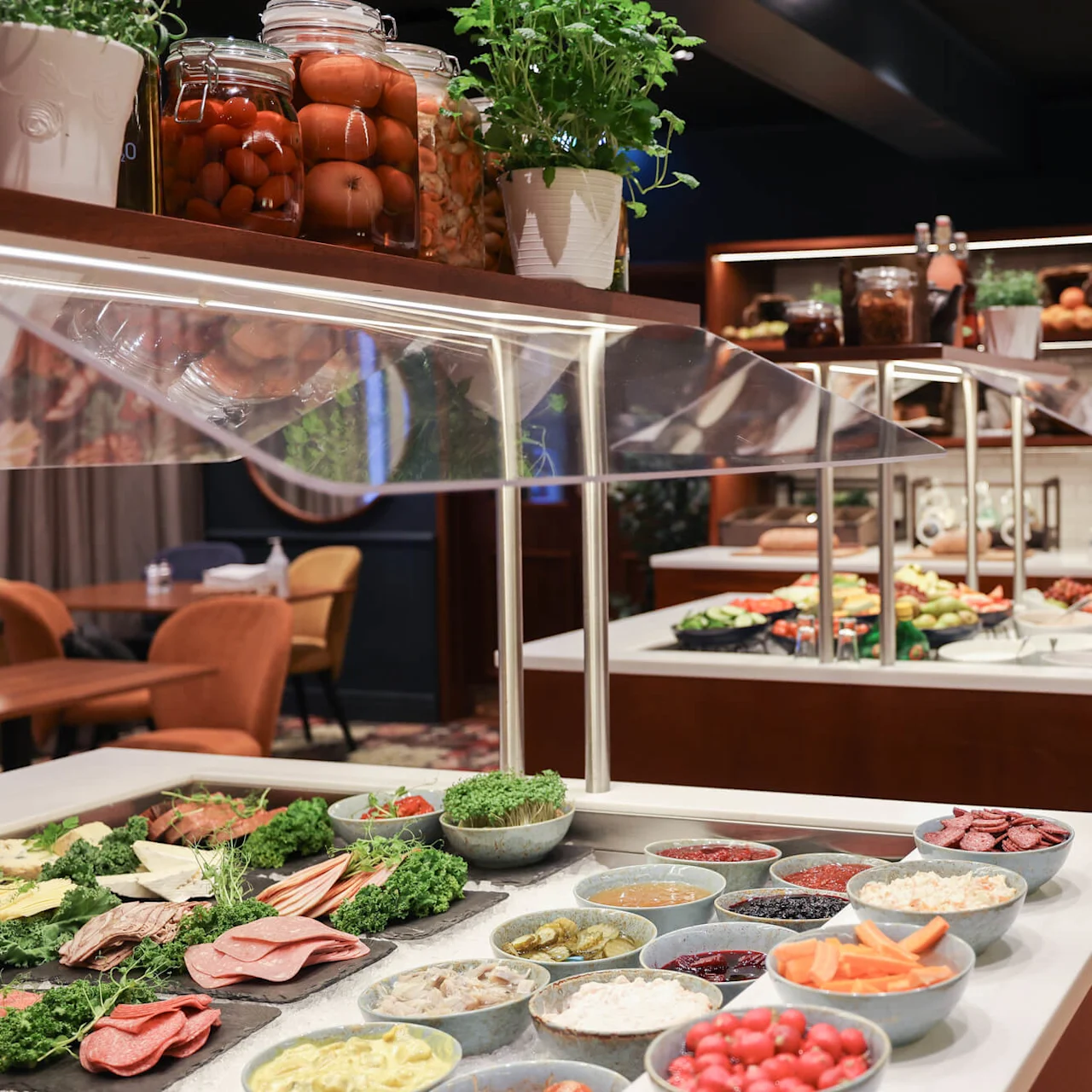 Buffet setup with assorted vegetables, meats, and garnishes arranged on a counter. Dining tables and chairs are visible in the background, creating a cozy restaurant atmosphere.