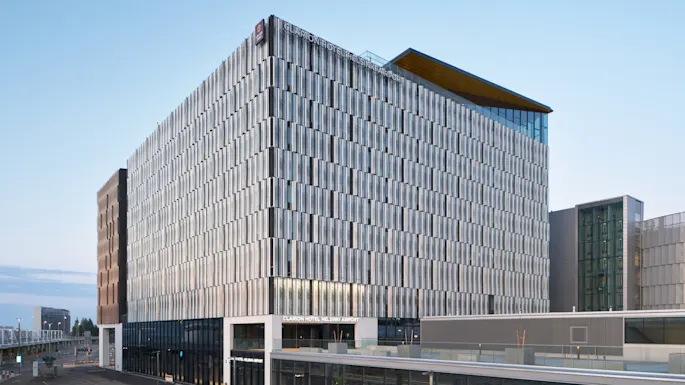 En moderne hotellbygning med glassfasade og skiltet "Clarion Hotel Helsinki Airport"; plassert ved siden av en jernbanestasjon. To parkerte biler og en klarblå himmel gir en rolig urban scene.
