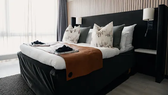 En seng, pænt redt med dekorative puder og et brunt tæppe, står i et moderne soveværelse omgivet af elegante væglamper og lyse gardiner, der filtrerer dagslys.