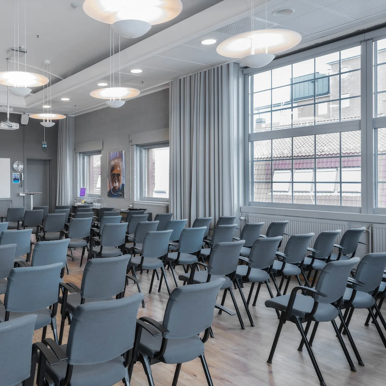 Rows of empty blue chairs face a large whiteboard, beneath bright lights. Windows span one wall, showing an urban backdrop, while a projector hangs from the ceiling.