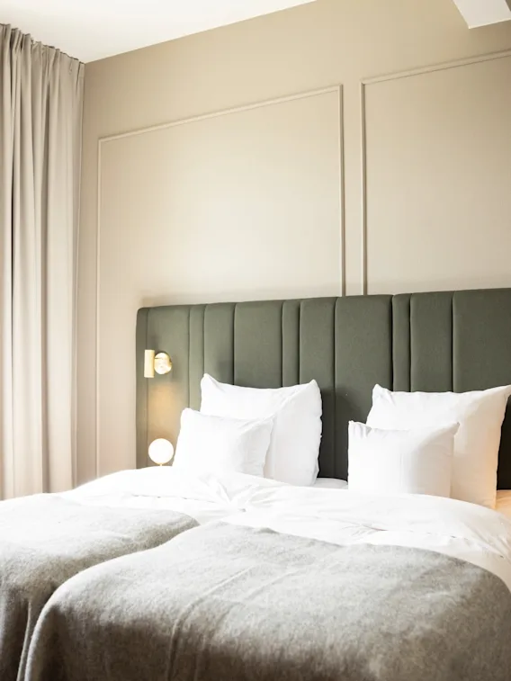 A bright hotel room with two comfortable beds, a dark green headboard, and a large window with light curtains.
