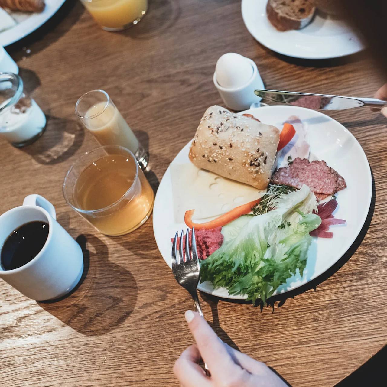 Morgenmadstallerken med en frørulle, skiver af skinke, salat og peberfrugt. Drikkevarer inklusive kaffe, juice og et blødkogt æg er på et træbord.