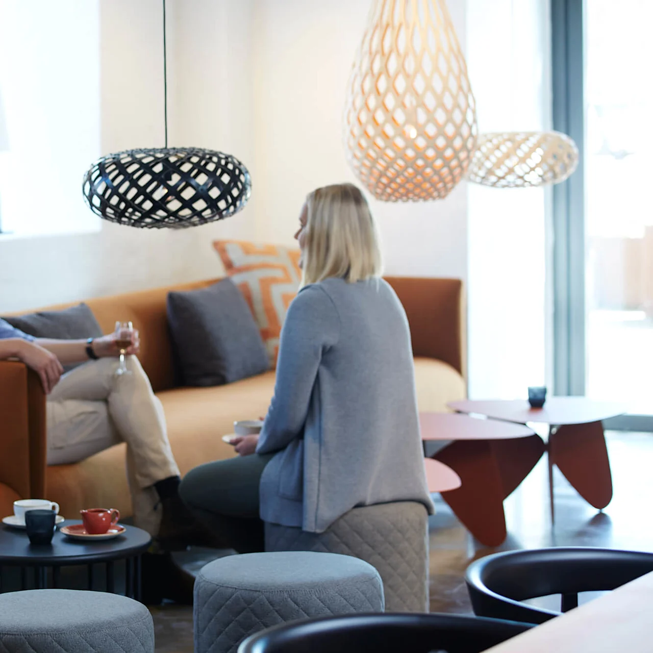 Two individuals conversing in a modern lounge with stylish hanging lamps and coffee tables.