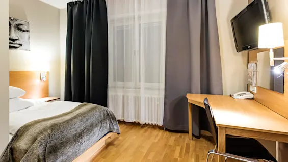 Et ryddig hotellrom med en seng med et pelspledd og et skrivebord med stol. En TV er montert på veggen over skrivebordet. Gardiner dekker et vindu.