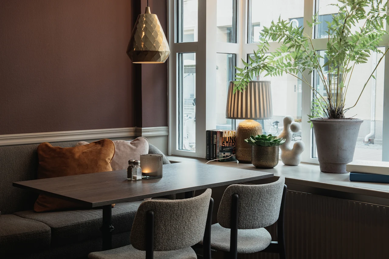 En mysig matplats med bord, stolar och en bänk vid ett fönster, dekorerad med växter och böcker inklusive MAN JUNG STED.