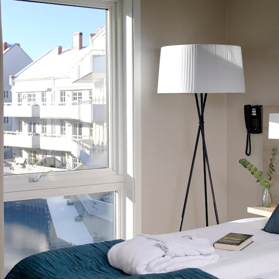 En prydligt bäddad hotellsäng med turkosa kuddar och en bok står vid ett stort fönster. En golvlampa och en telefon finns i närheten, med moderna byggnader utanför.