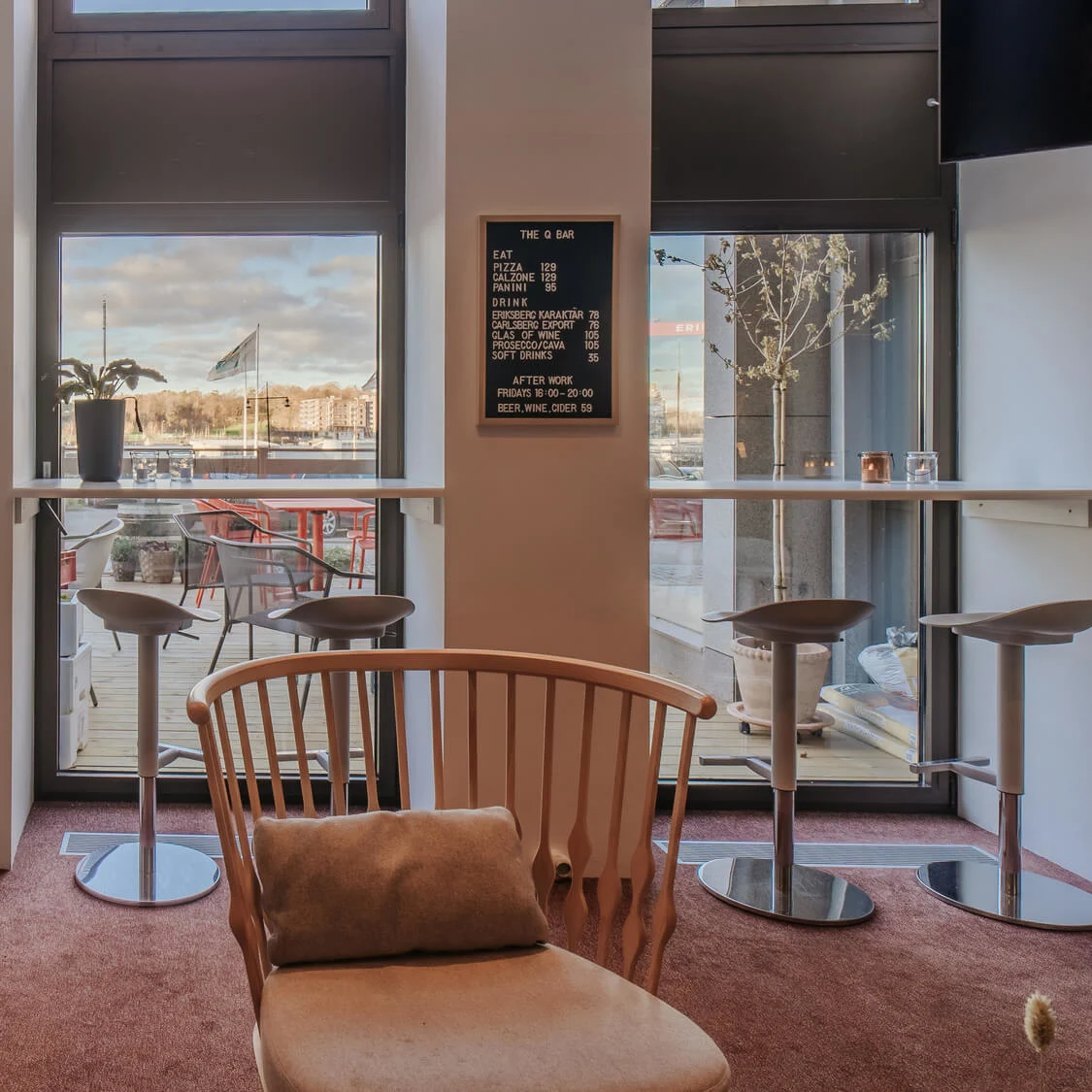 Tall potted plants sit on a carpet near windows with outdoor views and a high table with stools. A black menu board reads: "THE Q BAR," listing food and drinks.