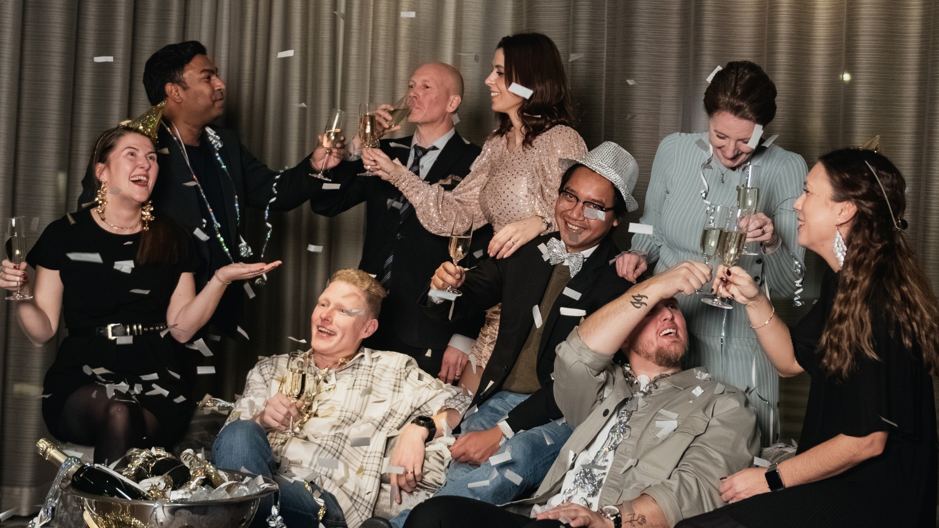 A group of people is celebrating, smiling, and holding champagne glasses, surrounded by confetti. They are dressed in party attire with a curtain backdrop.