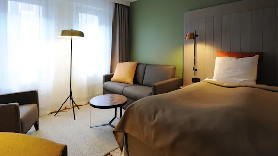 A cozy hotel room features a large bed with a wooden headboard, a sofa with cushions, and a floor lamp beside a window, creating a warm, inviting atmosphere.